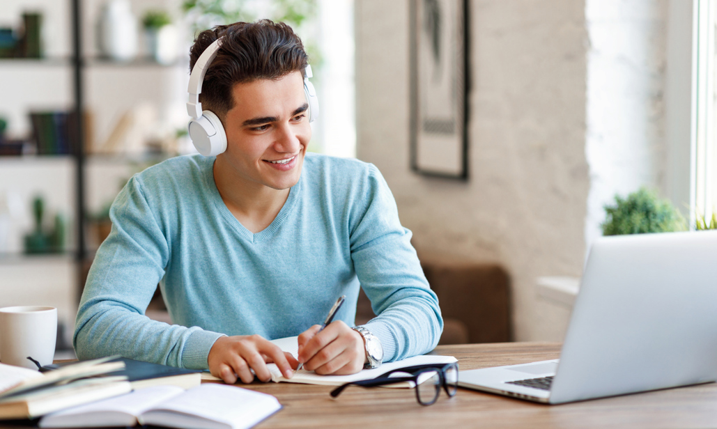 A man with headphones writing notes whilst looking at a laptop