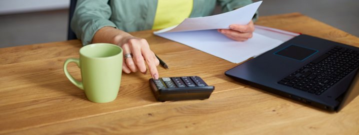 Picture of a hand tapping on a calculator with a laptop beside